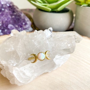 May include: A gold ring with three crescent moons and a white opal gemstone. The ring is displayed on a bed of clear quartz crystals.