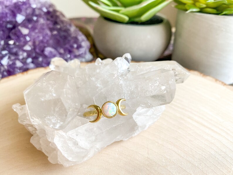 May include: A gold ring with three crescent moons and a white opal gemstone. The ring is displayed on a bed of clear quartz crystals.