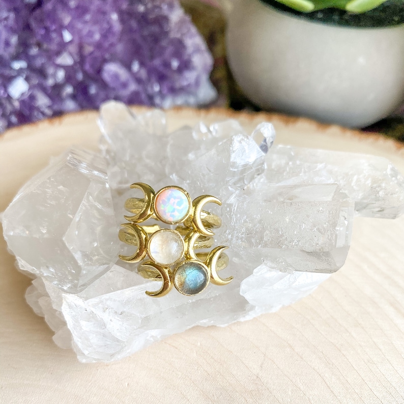 May include: Three gold rings with crescent moon designs and different colored gemstones. The top ring has a white opal, the middle ring has a white moonstone, and the bottom ring has a blue labradorite.