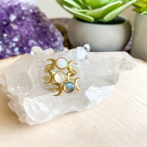 May include: Three gold rings with crescent moon designs and different colored stones. The stones are opal, moonstone, and labradorite. The rings are displayed on a cluster of clear quartz crystals.