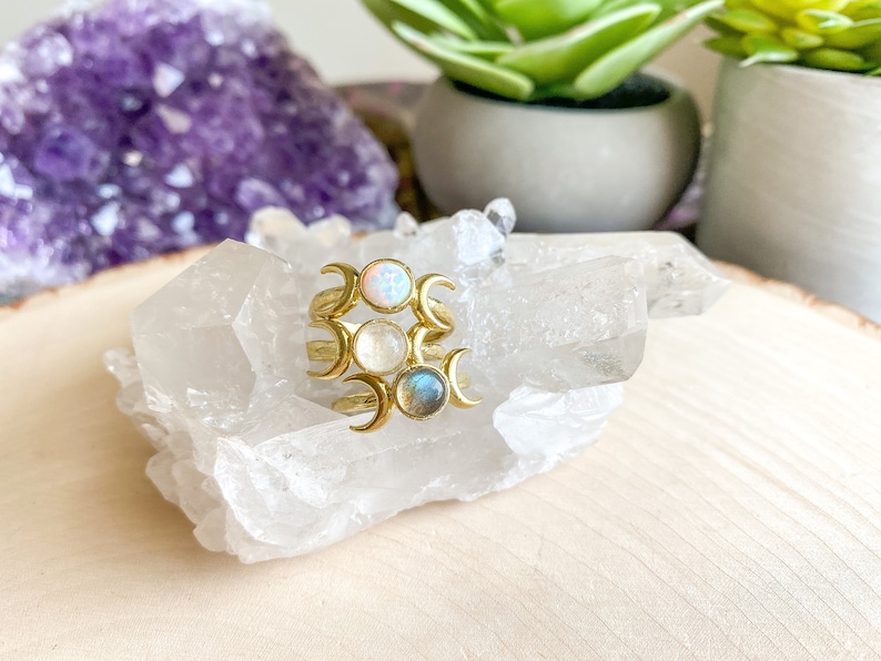 May include: Three gold rings with crescent moon designs and different colored stones. The stones are opal, moonstone, and labradorite. The rings are displayed on a cluster of clear quartz crystals.