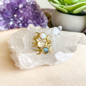 May include: Three gold rings with crescent moon designs and different colored stones. The stones are opal, moonstone, and labradorite. The rings are displayed on a white crystal.