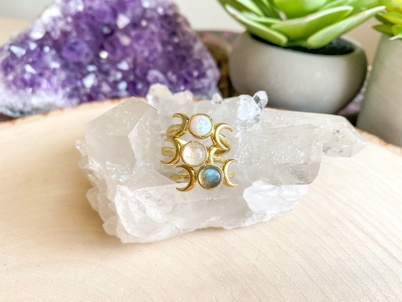 May include: Three gold rings with crescent moon designs and different colored stones. The stones are opal, moonstone, and labradorite. The rings are displayed on a white crystal.