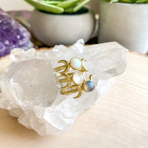 May include: Gold ring with three crescent moons and three gemstones: a white opal, a white moonstone, and a blue labradorite.