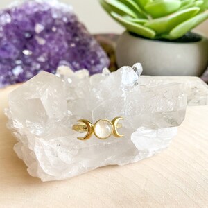 May include: A gold ring with three crescent moons and a white moonstone in the center, resting on a bed of white quartz crystals.