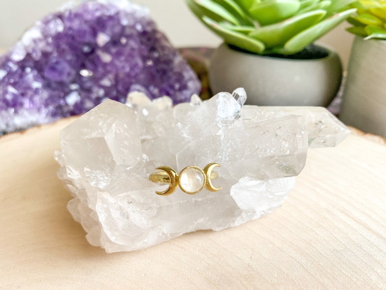 May include: A gold ring with three crescent moons and a white moonstone in the center, resting on a bed of white quartz crystals.