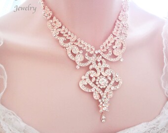 May include: Rose gold-toned necklace with intricate filigree and sparkling clear crystals. The necklace features a central pendant with a heart-shaped design and a delicate teardrop. The text "Queen Me Jewelry" is visible.
