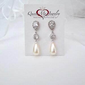 May include: Elegant drop earrings with a pear-shaped pearl and sparkling clear crystals. The earrings are silver-toned and feature a teardrop-shaped crystal at the top, a round crystal in the middle, and a cream-colored pearl at the bottom. The earrings are displayed on a white card with the text "Queen Me Jewelry".