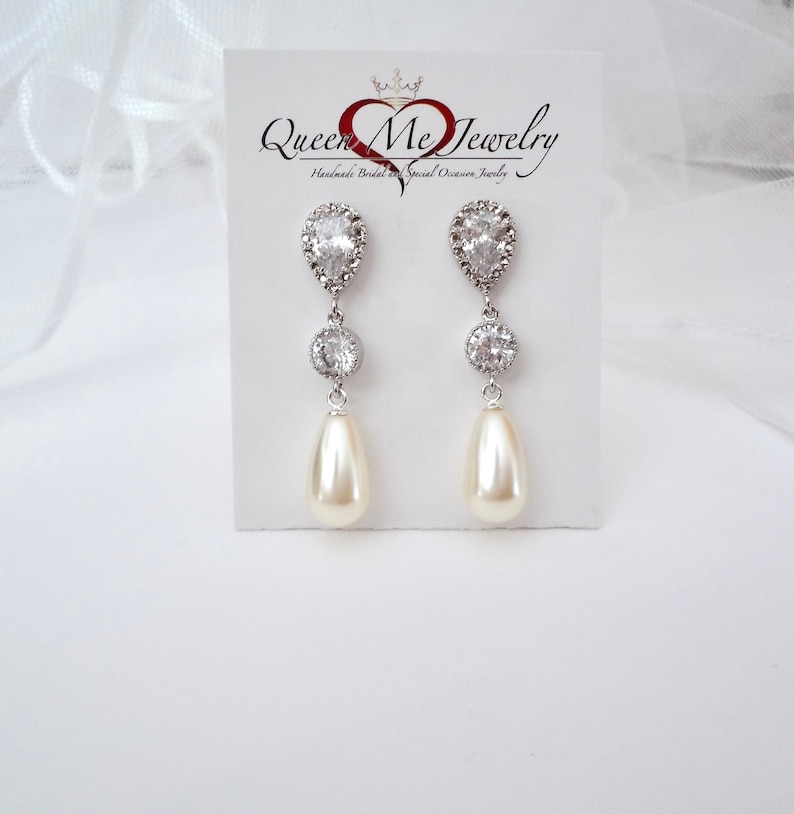 May include: Elegant drop earrings with a pear-shaped pearl and sparkling clear crystals. The earrings are silver-toned and feature a teardrop-shaped crystal at the top, a round crystal in the middle, and a cream-colored pearl at the bottom. The earrings are displayed on a white card with the text "Queen Me Jewelry".