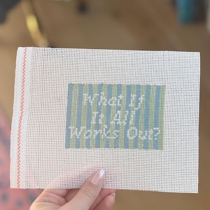 以下が含まれることがあります： 青と緑の「What If It All Works Out?」という文字が入った白いニードルポイントキャンバス。 キャンバスにはグリッドパターンがあり、左側に赤いボーダーがあります。 キャンバスは手で持たれています。