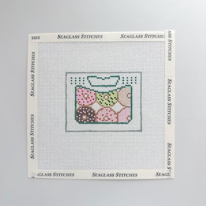 May include: A square needlepoint canvas featuring a central graphic of a rectangular bag filled with six colorful donuts in shades of pink, brown, and green. The text SEAGLASS STITCHES is printed repeatedly along the white borders of the canvas.