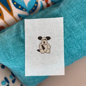 May include: A small, rectangular needlepoint canvas with a pixelated dog design. The dog has black ears and spots, a beige body, and a black tie. The canvas is white with a grid pattern, and is resting on a turquoise cushion.