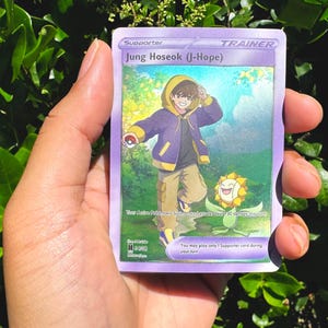May include: A purple Trainer card featuring Jung Hoseok (J-Hope) with a cartoon illustration of a person in a purple jacket and tan trousers. A smiling flower is also depicted. The card reads "Supporter" and includes game text.