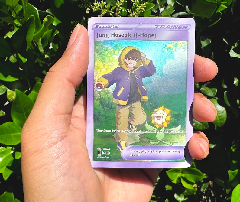 May include: A purple Trainer card featuring Jung Hoseok (J-Hope) with a cartoon illustration of a person in a purple jacket and tan trousers. A smiling flower is also depicted. The card reads "Supporter" and includes game text.