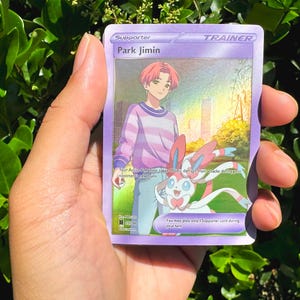 May include: A purple Trainer card featuring Park Jimin with pink hair and a striped shirt, alongside a Sylveon. The card has the text "Supporter" and "Trainer" at the top. The background shows a city and a sunny sky.