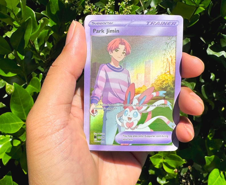 May include: A purple Trainer card featuring Park Jimin with pink hair and a striped shirt, alongside a Sylveon. The card has the text "Supporter" and "Trainer" at the top. The background shows a city and a sunny sky.