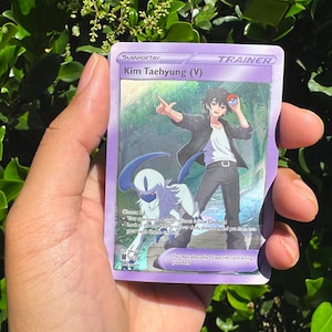 May include: A purple Trainer card featuring an illustration of Kim Taehyung (V). The card has a colourful background with a character holding a Pok&eacute; Ball. The card also features a blue and white creature.