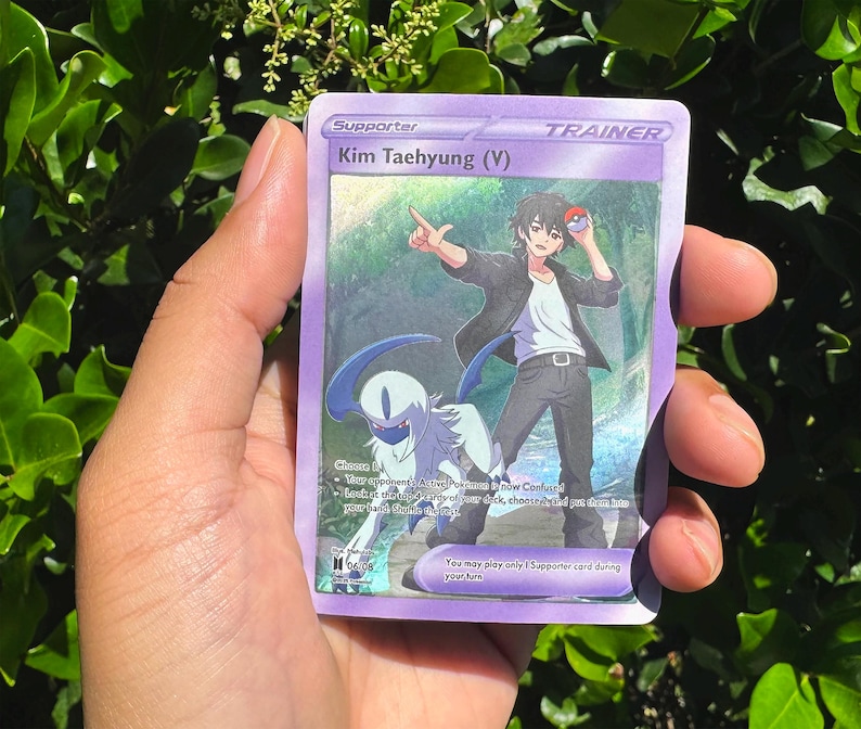 May include: A purple Trainer card featuring an illustration of Kim Taehyung (V). The card has a colourful background with a character holding a Pok&eacute; Ball. The card also features a blue and white creature.