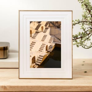 May include: Framed photograph of a stepwell with descending staircases. The image features a gold-colored frame and a white mat. The stepwell has beige walls and steps, with a dark pool of water at the bottom. A plant is visible in the background.