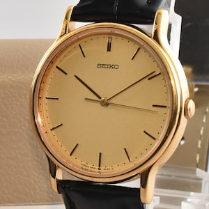 Op de afbeelding: Een goudkleurig Seiko polshorloge met een ronde champagnekleurige wijzerplaat, dunne zwarte wijzers en zwarte uurmarkeringen. Het horloge heeft een zwarte leren band met een textuur en wordt gepresenteerd naast een beige doos met het label JPgoldwatchstore.