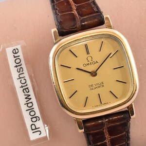 May include: A gold Omega De Ville Quartz watch with a rectangular face and brown leather band. The watch has a gold dial with black hands and markers. The watch is on a wrist.