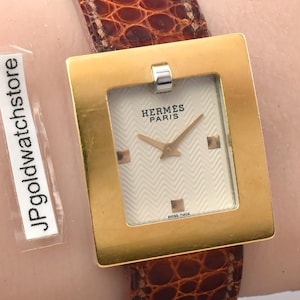 May include: A wristwatch with a square gold-tone metal case and a white textured dial featuring the text HERMES PARIS. The watch has a brown leather strap with a reptilian-like scale pattern. A small rectangular tag reads JPgoldwatchstore.