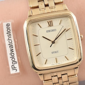 Könnte beinhalten: Eine goldfarbene Seiko Spirit Armbanduhr mit rechteckigem Zifferblatt und passendem Gliederarmband. Die Uhr hat ein cremefarbenes Zifferblatt mit schwarzen Zeigern und Markierungen. Die Uhr wird am Handgelenk getragen.
