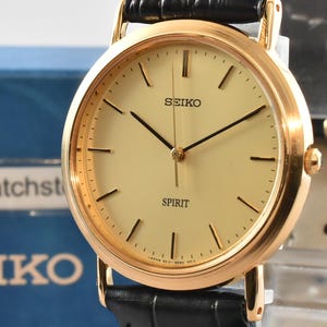 May include: A Seiko Spirit wristwatch featuring a round gold-tone case, a champagne-colored dial with thin black hour markers and hands, and a black leather strap with a textured finish. The watch is displayed in front of a blue Seiko box.