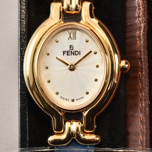 Puede incluir: Un reloj de pulsera Fendi dorado con esfera ovalada, fondo blanco y número romano en la posición de las doce. El reloj cuenta con una correa de cuero texturizado azul y se muestra junto a correas intercambiables en marrón, rojo, amarillo y naranja. Es visible una etiqueta con el texto JPgoldwatchstore.
