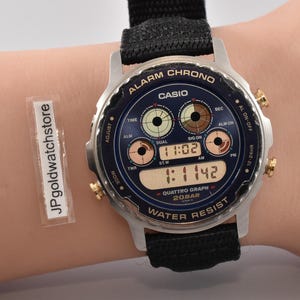 Op de afbeelding: Een Casio Alarm Chrono digitale horloge met een zwarte stoffen band. De wijzerplaat is donkerblauw met gouden accenten en geeft de tijd 11:02 en 1:11:42 weer. Het horloge is waterbestendig.