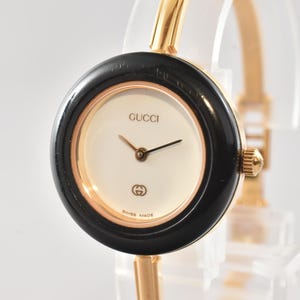 May include: A gold-tone bangle watch featuring a round case with a black bezel and a cream-colored dial. The dial displays the Gucci logo at the top, the double G emblem at the bottom, and the text SWISS MADE. It has gold-tone hands and a crown.