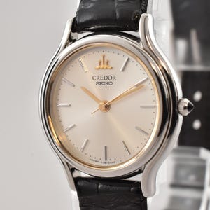 Op de afbeelding: Een Credor Seiko polshorloge met een ronde zilverkleurige wijzerplaat, goudkleurige wijzers en goudkleurige uurmarkeringen. Het horloge heeft een zilverkleurige lunette, een goudkleurige binnenring en een zwarte lederen band met een textuurafwerking.