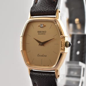 Puede incluir: Un reloj de pulsera Seiko Quartz Exceline con caja dorada en forma de tonel, esfera color champ&aacute;n con un diamante en la posici&oacute;n de las doce, manecillas doradas finas y una correa de cuero marr&oacute;n oscuro texturizada con corona dorada.