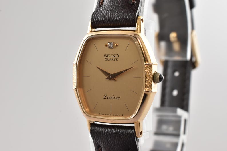 Puede incluir: Un reloj de pulsera Seiko Quartz Exceline con caja dorada en forma de tonel, esfera color champ&aacute;n con un diamante en la posici&oacute;n de las doce, manecillas doradas finas y una correa de cuero marr&oacute;n oscuro texturizada con corona dorada.