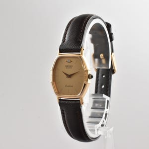 Puede incluir: Un reloj de pulsera Seiko Quartz Exceline con caja dorada en forma de tonel, un diamante en la posici&oacute;n de las doce, esfera dorada y correa de cuero marr&oacute;n oscuro con hebilla y herrajes de color dorado a juego.