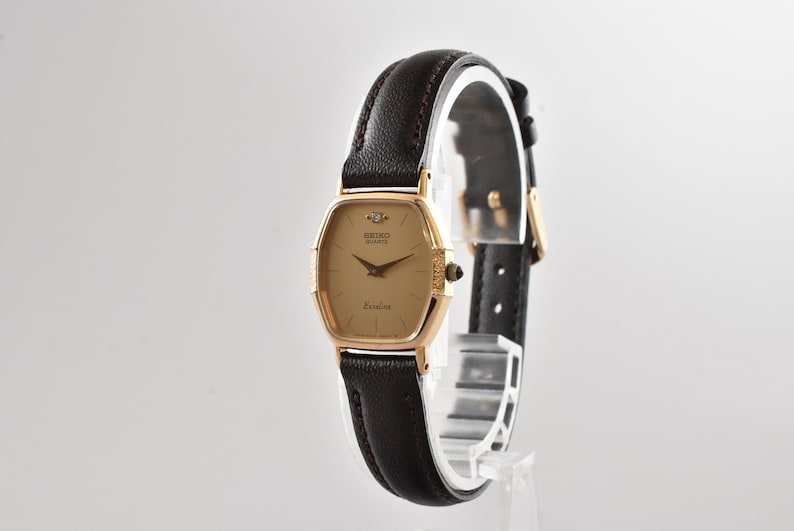 Puede incluir: Un reloj de pulsera Seiko Quartz Exceline con caja dorada en forma de tonel, un diamante en la posici&oacute;n de las doce, esfera dorada y correa de cuero marr&oacute;n oscuro con hebilla y herrajes de color dorado a juego.