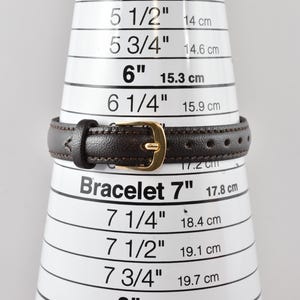 Puede incluir: Una correa de reloj de cuero marr&oacute;n oscuro con una hebilla de metal dorado, colocada sobre un cono de medici&oacute;n blanco que indica una medida de 17,8 cm. La correa presenta costuras visibles en los bordes y varios orificios de ajuste.