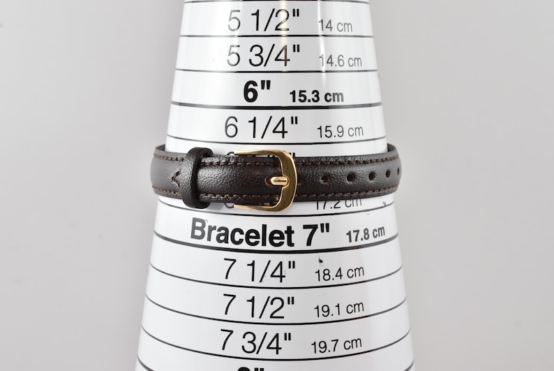 Puede incluir: Una correa de reloj de cuero marr&oacute;n oscuro con una hebilla de metal dorado, colocada sobre un cono de medici&oacute;n blanco que indica una medida de 17,8 cm. La correa presenta costuras visibles en los bordes y varios orificios de ajuste.