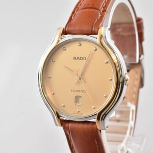 Op de afbeelding: Een Rado Florence polshorloge met een ronde goudkleurige wijzerplaat, stipvormige uurmarkeringen, gouden wijzers en een datumvenster op de zes-uur-positie. Het horloge heeft een tweekleurige zilveren en gouden lunette en een bruine leren band met stiksels.