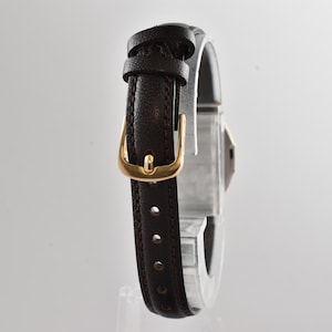 Puede incluir: Una correa de reloj de cuero marr&oacute;n oscuro con una superficie texturizada y costuras visibles a lo largo de los bordes. La correa cuenta con una hebilla de metal en tono dorado pulido y dos trabillas de cuero a juego.