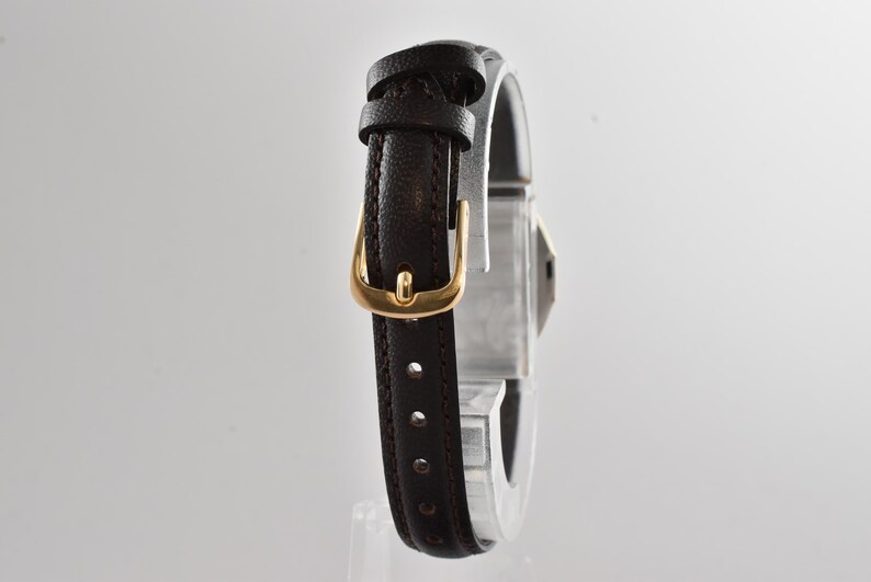 Puede incluir: Una correa de reloj de cuero marr&oacute;n oscuro con una superficie texturizada y costuras visibles a lo largo de los bordes. La correa cuenta con una hebilla de metal en tono dorado pulido y dos trabillas de cuero a juego.