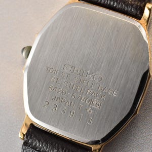 Puede incluir: Un reloj de pulsera Seiko en tono dorado con correa de cuero negro. La tapa trasera de acero inoxidable tiene grabado: SEIKO, 10K ST. STEEL BASE, ST. STEEL BACK, 8420-5780, JAPAN T y el n&uacute;mero de serie 233913. Acabado de metal cepillado.