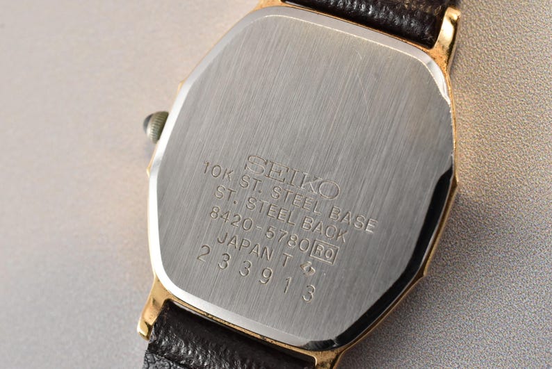 Puede incluir: Un reloj de pulsera Seiko en tono dorado con correa de cuero negro. La tapa trasera de acero inoxidable tiene grabado: SEIKO, 10K ST. STEEL BASE, ST. STEEL BACK, 8420-5780, JAPAN T y el n&uacute;mero de serie 233913. Acabado de metal cepillado.