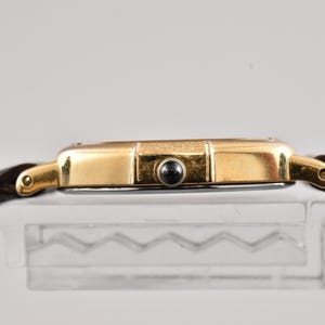 Puede incluir: Una caja de reloj en tono dorado con una correa de cuero marr&oacute;n oscuro. El perfil lateral muestra un acabado dorado met&aacute;lico, una peque&ntilde;a corona cabochon negra y asas curvas que conectan con la correa. La caja tiene una superficie pulida.