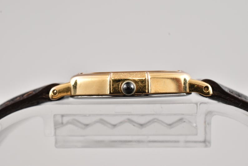 Puede incluir: Una caja de reloj en tono dorado con una correa de cuero marr&oacute;n oscuro. El perfil lateral muestra un acabado dorado met&aacute;lico, una peque&ntilde;a corona cabochon negra y asas curvas que conectan con la correa. La caja tiene una superficie pulida.