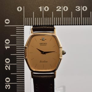 Puede incluir: Un reloj de pulsera Seiko Quartz Exceline en tono dorado con esfera color champ&aacute;n, un marcador de diamante en la posici&oacute;n de las 12 y correa de cuero marr&oacute;n oscuro, junto a una regla negra en forma de L que muestra 2,5 cm de ancho.