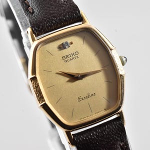 Puede incluir: Un reloj de pulsera Seiko Quartz Exceline con caja hexagonal en tono dorado, esfera dorada texturizada con un diamante en la posici&oacute;n de las doce, manecillas doradas finas y correa de cuero marr&oacute;n oscuro con costuras visibles.
