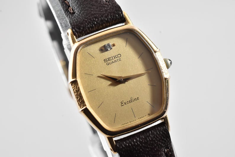 Puede incluir: Un reloj de pulsera Seiko Quartz Exceline con caja hexagonal en tono dorado, esfera dorada texturizada con un diamante en la posici&oacute;n de las doce, manecillas doradas finas y correa de cuero marr&oacute;n oscuro con costuras visibles.