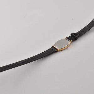 Puede incluir: Un reloj de pulsera con una correa negra delgada y una caja de tono dorado con una placa trasera de color plateado. El reloj incluye un cierre de hebilla de tono dorado y una serie de orificios de ajuste a lo largo de la banda negra.