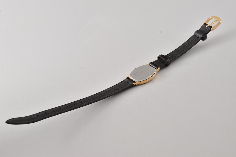 Puede incluir: Un reloj de pulsera con una correa negra delgada y una caja de tono dorado con una placa trasera de color plateado. El reloj incluye un cierre de hebilla de tono dorado y una serie de orificios de ajuste a lo largo de la banda negra.